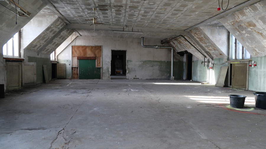 Blick in den Dachbodenraum der Lausitzer Str. 10, in dem der Hauptteil der Ausstellung stattfindet, ein leerer heller Raum (3 Fenster sichtbar) mit gegenüberliegender Türoffnung und rechts Türöffnung zu einem Seitenraum